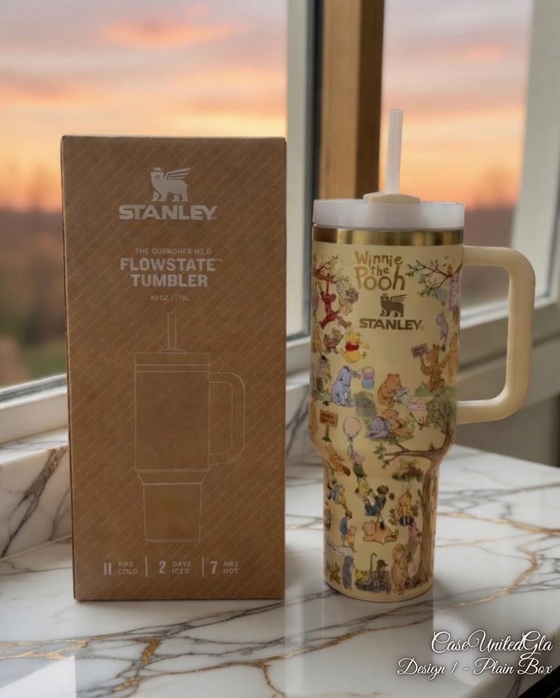 May include: A beige Stanley Flowstate tumbler with a Winnie the Pooh design, a white straw, and a handle. The tumbler is next to its cardboard box. The box has the Stanley logo and product information. The tumbler holds 40 oz.