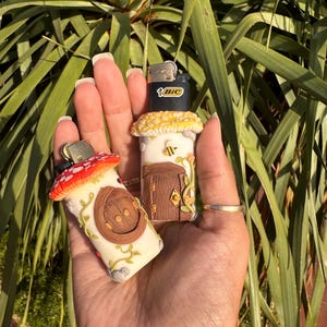 Pode incluir: Dois isqueiros BIC decorados com argila polimérica para se assemelharem a casas de cogumelos em miniatura. Um apresenta um telhado de cogumelo vermelho com uma porta arqueada castanha, o outro um telhado amarelo, porta castanha e uma abelha.