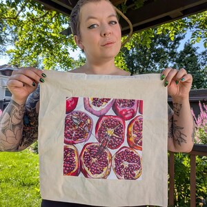 Op de afbeelding: Een beige katoenen draagtas met een vierkante grafische print van meerdere gehalveerde granaatappels met dieprode zaden en een klein insect dat op een van de vruchtstukken rust, gecentreerd op de voorzijde van de tas.