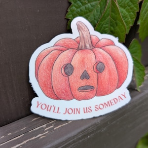 May include: A sticker featuring a red-orange pumpkin with a carved face and a stem. The text "YOU'LL JOIN US SOMEDAY" is printed below the pumpkin. The sticker is on a wooden surface with green leaves in the background.