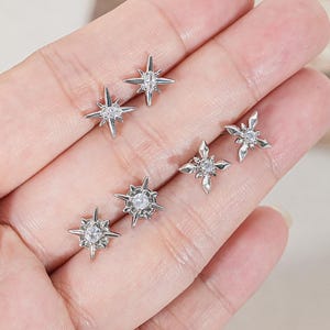 May include: A set of five silver-toned stud earrings, each featuring a multi-pointed star design with a central clear stone. The earrings vary slightly in their starburst patterns, with some having elongated points and others having a more compact shape.