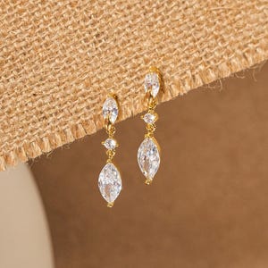 May include: A pair of gold-colored drop earrings with clear, marquise-cut cubic zirconia stones. The earrings have a delicate design with three stones per earring, creating a sparkling and elegant look. The earrings are displayed against a neutral background.