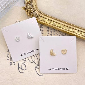 May include: Two pairs of stud earrings on white cards. One pair has a pretzel and a croissant, the other a pretzel and a croissant. The cards have the text "THANK YOU" and paw prints.