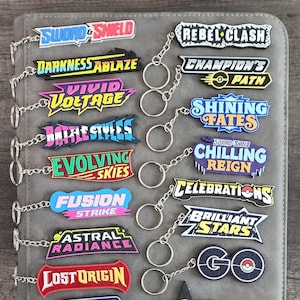 May include: A selection of vibrant keyrings with text designs, including "Sword & Shield", "Darkness Ablaze", and "Vivid Voltage". Each keyring is attached to a silver chain and ring. The keyrings are on a grey surface.