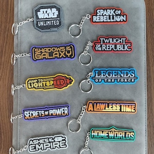 Peut inclure: Plusieurs porte-clés sur le thème de Star Wars, avec des titres tels que "Star Wars Unlimited", "Spark of Rebellion" et "Shadows of the Galaxy". Chaque porte-clés est doté d'une chaîne et d'un anneau argentés, présentés sur un porte-cartes gris.