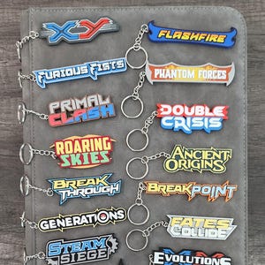 Puede incluir: Una colección de llaveros coloridos con varios diseños de texto, incluyendo "FLASHFIRE" y "EVOLUTIONS." Los llaveros están sujetos a un tarjetero gris, con temática de cartas coleccionables.