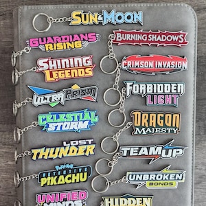 Può includere: Una collezione di portachiavi colorati con titoli come "Sun & Moon", "Guardians Rising" e "Burning Shadows". Ogni portachiavi ha un anello e una catena argentati, presentati su una superficie grigia. I portachiavi sono un mix di colori, tra cui rosa, blu e giallo.