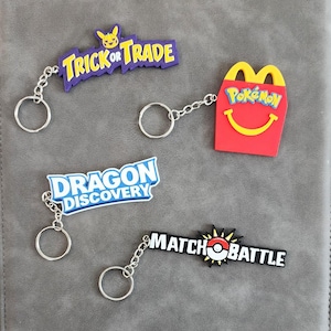 Può includere: Quattro portachiavi colorati con catene e anelli argentati. I portachiavi presentano le scritte "Trick or Trade", "Dragon Discovery", "Match Battle" e una scatola di patatine fritte rossa con il logo Pokémon.