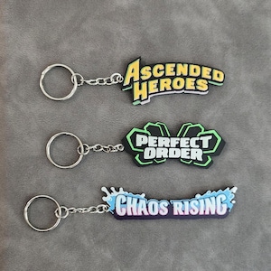 May include: Three keychains with metal rings and chains. The top keychain features yellow text reading ASCENDED HEROES. The middle keychain has a black and green shape with white text reading PERFECT ORDER. The bottom one has blue splash accents and white text reading CHAOS RISING.