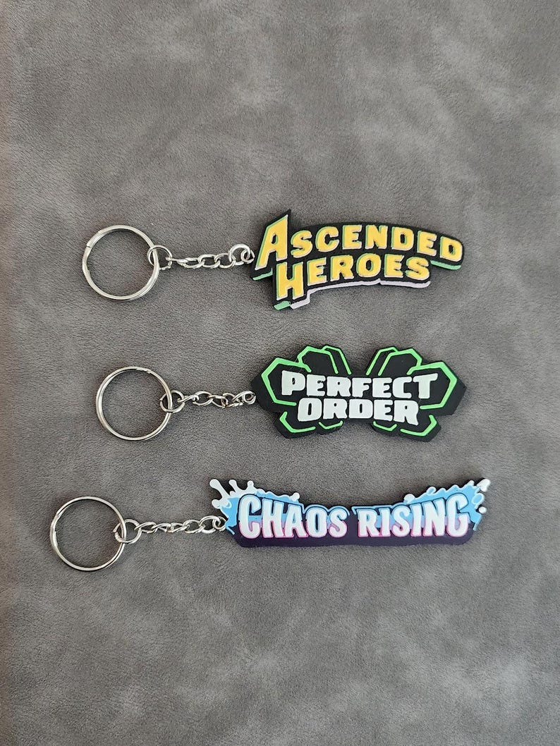 May include: Three keychains with metal rings and chains. The top keychain features yellow text reading ASCENDED HEROES. The middle keychain has a black and green shape with white text reading PERFECT ORDER. The bottom one has blue splash accents and white text reading CHAOS RISING.