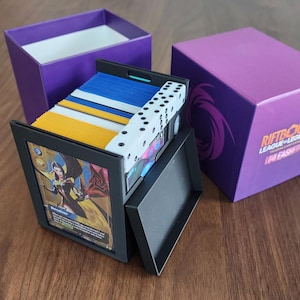 May include: A black plastic trading card storage box containing stacks of blue and yellow cards, with a white six-sided die on top. The front displays a character card labeled Deceiver. A purple box with the text RIFTBOUND LEAGUE OF LEGENDS is nearby.