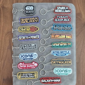 May include: A collection of seventeen Star Wars Unlimited themed keychains featuring various set and expansion logos including Spark of Rebellion, Shadows of the Galaxy, Twilight of the Republic, Legends of the Force, and others in diverse colours.