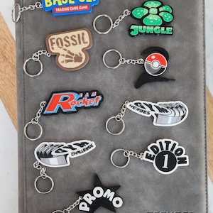 May include: A selection of keyrings featuring designs and text such as "BASE SET", "FOSSIL", "JUNGLE", "Rocket", "CHALLENGE", "PROMO", "HEROES", and "EDITION 1". The keyrings are displayed on a grey binder.