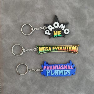 May include: Three keychains with silver metal rings and chains. The top is a black star with white text reading PROMO and yellow and green text reading ME. The middle is a long shape with yellow text MEGA EVOLUTION. The bottom is blue with pink and light blue text PHANTASMAL FLAMES.