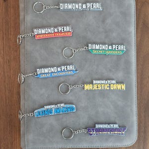 May include: A gray binder with silver keychains attached. Each keychain has a colorful rectangular charm with the words "Diamond & Pearl" and a different title, including "Mysterious Treasures" and "Secret Wonders."