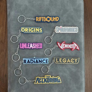 May include: A collection of seven rectangular keychains with silver metal rings, each featuring different text designs: RIFTBOUND, ORIGINS, SPIRITFORGED, UNLEASHED, VENDETTA, RADIANCE, LEGACY, and THE RECKONING, displayed in various colors and fonts.