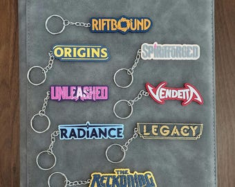 May include: A collection of seven rectangular keychains with silver metal rings, each featuring different text designs: RIFTBOUND, ORIGINS, SPIRITFORGED, UNLEASHED, VENDETTA, RADIANCE, LEGACY, and THE RECKONING, displayed in various colors and fonts.