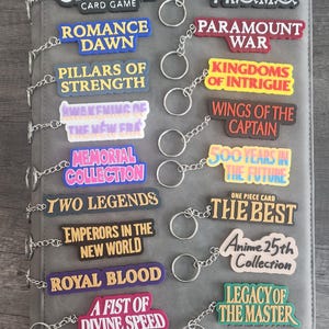Puede incluir: Colección de llaveros coloridos con textos del juego de cartas "One Piece", incluyendo "Promo", "Paramount War" y "Legacy of the Master". Los llaveros están sujetos a una carpeta gris.