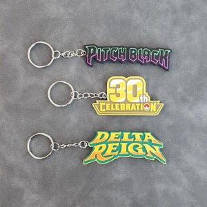 May include: Three metal keychains with attached rubberized charms. The top charm is black with pink text reading PITCH BLACK. The middle charm is yellow and white with text reading 30th CELEBRATION. The bottom charm is green with yellow text reading DELTA REIGN.