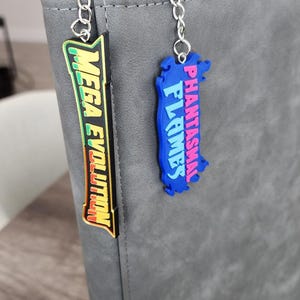 May include: A gray zippered binder featuring two hanging keychains. One keychain is black with green and yellow text reading MEGA EVOLUTION. The second keychain is blue with pink text reading PHANTASMAL FLAMES. Both are attached by silver metal chains.