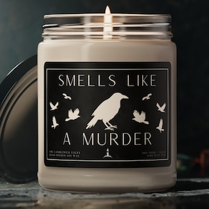 May include: A lit candle in a glass jar with a silver lid. The label reads "Smells Like A Murder" with a raven and flying birds. The candle is on a stone surface.