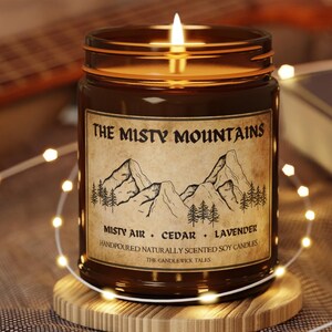 Pode incluir: Um frasco de vela de vidro castanho com um rótulo que diz "The Misty Mountains" e "Misty Air, Cedar, Lavender". A vela é feita à mão e feita com cera de soja natural.