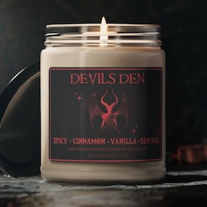 Könnte beinhalten: Ein schwarzes und rotes Kerzengefäß mit einem Etikett, auf dem "Devils Den" und "Spicy - Cinnamon - Vanilla - Sensual" steht. Die Kerze ist handgegossen und natürlich duftend.