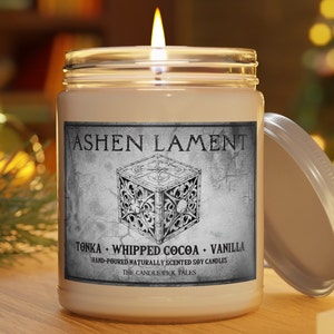 May include: A glass jar candle with a white label that reads "ASHEN LAMENT" and "TONKA - WHIPPED COCOA - VANILLA" with a decorative image of a box. The label also reads "HAND-POURED NATURALLY SCENTED SOY CANDLES" and "THE CANDLEWICK TALES".