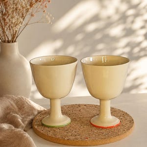 Può includere: Due calici in ceramica color crema con coppe lisce e arrotondate e steli robusti. Un calice presenta un sottile bordo verde alla base, mentre l'altro ha un sottile bordo arancione. Ogni calice è contrassegnato da un piccolo numero bianco.