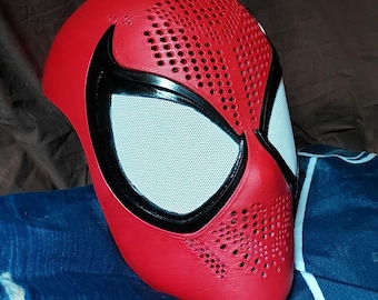 Puede incluir: Una máscara facial completa de color rojo con grandes lentes oculares blancas cubiertas de malla y delineadas en negro. La superficie de la máscara tiene una textura con un patrón de pequeños orificios de ventilación circulares.