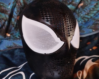 May include: A black full-face mask featuring a textured surface with small circular perforations across the forehead and chin areas. The mask includes two large, white, mesh-covered eye lenses that are shaped with sharp, angular outer edges.