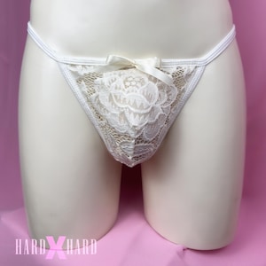 May include: Ivory lace thong underwear with a floral design on the front. The thong features a small bow at the waist and a delicate, see-through lace pattern. The underwear is displayed on a mannequin against a pink background.