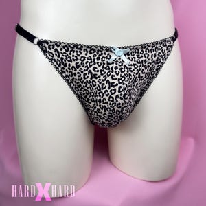 Peut inclure: String léopard avec bordure noire et un petit nœud bleu clair. Le sous-vêtement est présenté sur un mannequin. Le tissu présente un motif de taches noires sur fond crème. Le string a une fine lanière noire de chaque côté.
