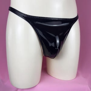 May include: Black thong underwear with a shiny, patent leather-like finish. The garment has a thin waistband and a contoured pouch. The edges are trimmed with a textured, stitched detail. The underwear is displayed on a mannequin.