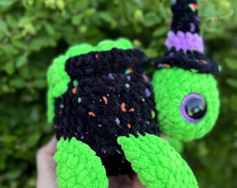 May include: A handmade crochet turtle in bright green and black, with a witch hat. The turtle's shell is black with colorful flecks, and the hat is black with a purple band. The turtle has large, round eyes.