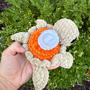 Könnte beinhalten: Ein handgefertigtes Häkeltier-Schildkrötenplüschtier. Der Kopf, die Flossen und die Beine der Schildkröte sind beige. Der Panzer ist orange mit einem weißen und blauen Blumenmuster. Das Plüschtier wird in einer Hand gehalten.