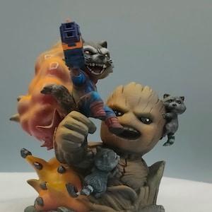 May include: A brown and green figurine of Groot from Guardians of the Galaxy, holding a raccoon figurine with a gun. The raccoon is wearing a red and blue costume. The figurine is set on a base with a brown and orange textured surface.