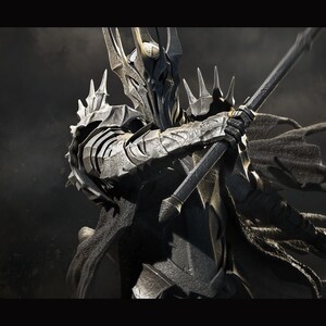 May include: A dark gray and silver action figure of a warrior wearing a helmet with a horned crown and a flowing cape. The figure is holding a spear in its right hand and is posed in a fighting stance. The figure is set against a dark gray background with clouds.