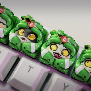 May include: Four custom keycaps featuring a cartoon Medusa design with green snakes, white skin, and yellow eyes. The keycaps are on a keyboard with white keys. The Nom Nom Figures logo is in the upper right corner.