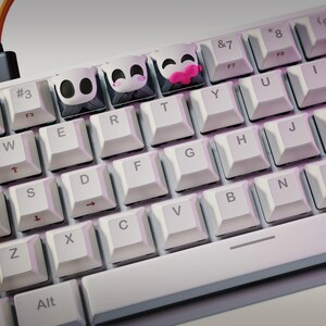 May include: A white mechanical keyboard with custom keycaps featuring panda and heart designs. The keyboard has a yellow shift key and a black cable. The logo "nom nom Figures" is in the top left corner.
