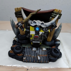 Puede incluir: Una figura de robot amarilla y negra con un ojo azul, sentada en un trono hecho de neumáticos y madera. El robot sostiene un palo en su mano derecha y una pistola en su mano izquierda. El trono está decorado con una tela marrón y una cuerda.