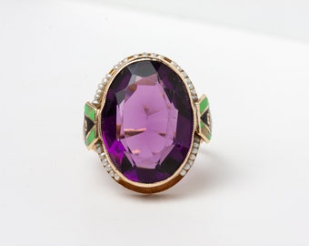 Puede incluir: Un anillo de oro con una gran piedra preciosa ovalada de color púrpura. El anillo está adornado con pequeñas perlas y detalles de esmalte verde y negro. Una joya de estilo vintage.
