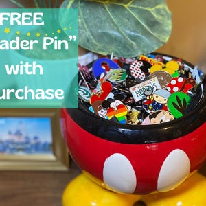 May include: A red, black, and white Mickey Mouse-themed bowl overflowing with colorful enamel pins. The pins feature various designs and characters. Text in the image reads "FREE Trader Pin with Purchase."