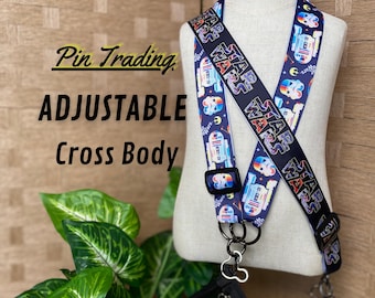 May include: A Star Wars themed adjustable cross body pin trading strap featuring a dark blue background with repeating colorful droid and helmet graphics. The strap includes a black plastic slider and metal hardware attached to two black pouches.