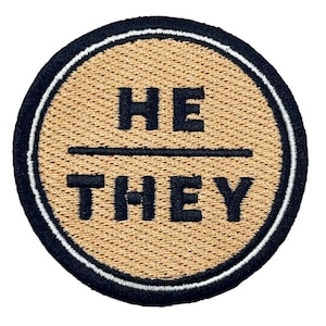 Könnte beinhalten: Ein runder, brauner gestickter Aufnäher mit schwarzem Rand und weißem Umriss. Der Aufnäher trägt die Aufschrift "HE/THEY" in Schwarz.