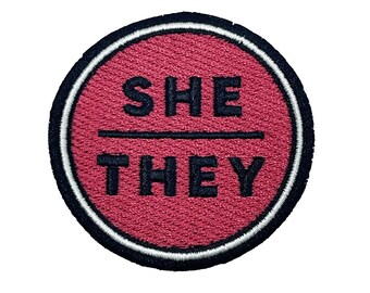 May include: A red circular embroidered patch with a white border and black stitching. The text "SHE / THEY" is embroidered in black.