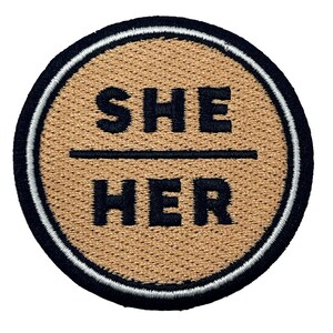 Può includere: Un cerotto rotondo in tessuto marrone con ricamo nero. Il testo "SHE / HER" è ricamato in nero sul cerotto.