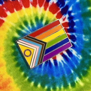 May include: A rainbow pride flag pin with a yellow triangle and a circle in the centre. The pin has a black, brown, light pink, white, light blue, dark blue, purple, green, yellow, and red stripe pattern.