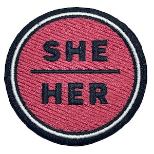Könnte beinhalten: Ein runder, rosafarbener Stoff-Patch mit weißem Rand und schwarzen Nähten. Der Text "SHE / HER" ist in Schwarz gestickt.