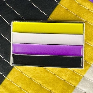 Peut inclure: Une épingle en métal aux couleurs du drapeau de la fierté non binaire. Le drapeau a quatre bandes horizontales : jaune, blanc, violet et noir.